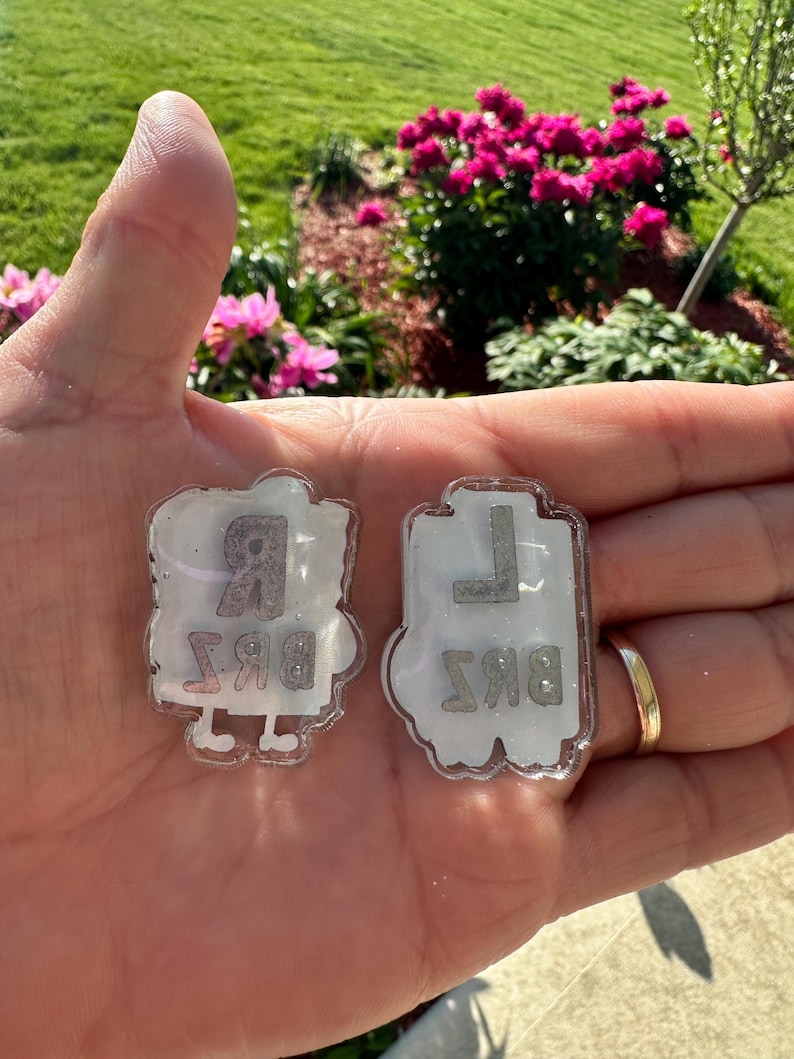 May include: Two white, semi-translucent acrylic charms with glitter accents. The left charm features the letter R above the letters BRZ. The right charm features the letter L above the letters BKS. Both charms have small white feet at the bottom.