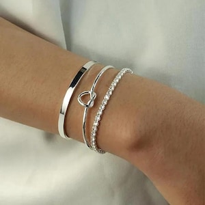 May include: Three silver bracelets, one with a knot design, one with a thin band, and one with a row of small rhinestones.