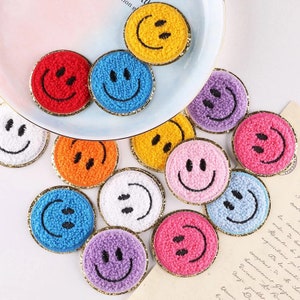 May include: A selection of colourful, round smiley face patches. The patches are in various colours, including red, blue, yellow, orange, white, purple, pink, and light blue. Each patch has a black embroidered smiley face and a gold trim.