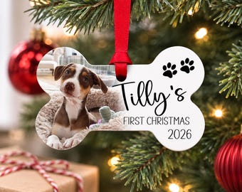 Könnte beinhalten: Ein knochenförmiger, weißer Weihnachtsschmuck, der an einem roten Band hängt. Die linke Seite zeigt ein Foto eines braun-weißen Welpen. Die rechte Seite enthält den schwarzen Text "Tilly's FIRST CHRISTMAS 2026" sowie zwei schwarze Pfotenabdruck-Symbole.