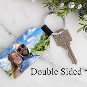 May include: Double-sided leather keychain featuring a photo of a couple against a blue sky and green trees. The keychain has a silver key ring and a black leather strap.