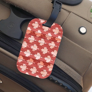 May include: A red and white luggage tag with a maple leaf pattern. The tag is attached to a brown suitcase.