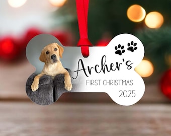 May include: A bone-shaped Christmas ornament featuring a photo of a golden Labrador puppy. The ornament has the text "Archer's First Christmas 2025" and paw prints. It is suspended by a red ribbon.