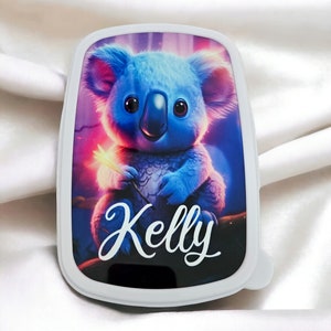 Könnte beinhalten: Weiße Lunchbox mit einem bunten Bild eines blauen Koalas, der einen leuchtenden Stock hält. Die Lunchbox trägt den Namen "Kelly" in weißer Kursivschrift auf der Vorderseite.