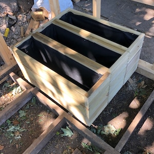 May include: A wooden planter box with three compartments, each lined with black fabric. The planter is sitting on a wooden platform.