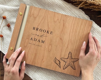 May include: A rectangular wooden photo album with a light brown grain, featuring the engraved text BROOKE and ADAM with the date 09.20.2025. The cover includes a detailed etched illustration of a starfish and a seashell, secured by gold metal screws.