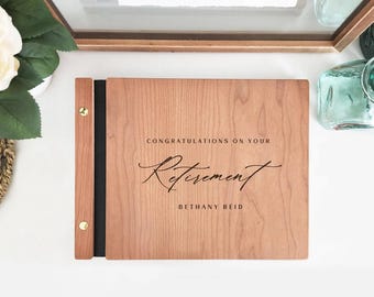 May include: A rectangular wooden photo album with a light brown wood grain finish. The cover features the black text CONGRATULATIONS ON YOUR RETIREMENT and the name BETHANY REID. A black spine is secured with two gold-colored metal binding screws.