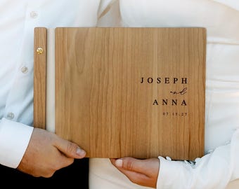 May include: A rectangular wooden photo album with a light brown grain finish. The cover features the black printed text JOSEPH and ANNA with the date 07.15.27 below. A vertical wooden spine strip is attached with a gold-colored metal screw post.