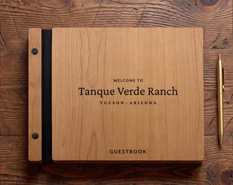 Peut inclure: Un livre d'or rectangulaire en bois avec une finition grainée brun clair, doté d'une reliure noire fixée par deux vis noires. La couverture porte l'inscription gravée "WELCOME TO Tanque Verde Ranch TUCSON - ARIZONA GUESTBOOK". Un stylo doré est posé à droite.