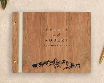 Peut inclure: Un album photo rectangulaire en bois avec une finition en grain de bois brun clair. La couverture présente le texte gravé AMELIA AND ROBERT, la date NOVEMBER 21, 2027, et un graphique de montagne noir. Il a une reliure blanche et deux vis dorées.