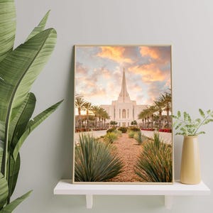 May include: A rectangular framed wall art print featuring a photograph of a white temple with a tall central spire, surrounded by palm trees and desert landscaping under a soft orange and blue sunset sky, displayed on a white shelf.