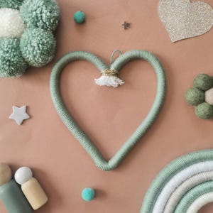 May include: A light green yarn heart-shaped wall decoration, with a gold ring and white tassel. The image includes a rainbow, pom-poms, stars, and other decorative elements in green, white, and beige, creating a whimsical aesthetic.