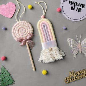 May include: Handmade macrame ornaments, featuring a pink and white lollipop with a bow, and a rainbow-coloured gnome with a white beard. The image also includes a pink heart, a butterfly, and the text "Macrame Be Me".