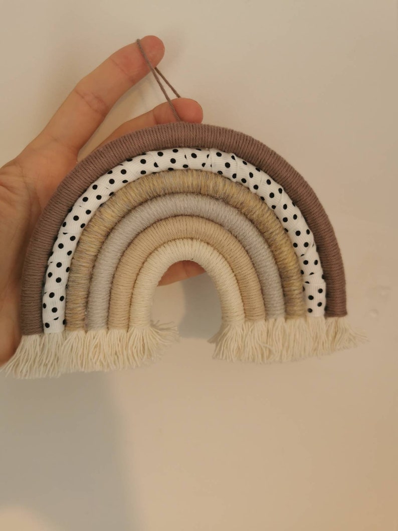 May include: A brown and white polka dot fabric rainbow wall hanging with beige and cream yarn strands. The rainbow has a fringe at the bottom.