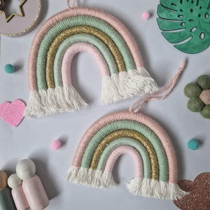May include: Two handmade fibre art rainbow wall hangings featuring wrapped yarn in pastel pink, sage green, and metallic gold thread. Each rainbow has white fringe at both ends and a matching pink yarn loop for hanging on a wall or surface.