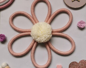 May include: A handmade, pink yarn flower wall hanging with a cream-colored pom-pom center. The flower is surrounded by decorative elements, including a small rainbow, wooden star, and paper flowers. The overall aesthetic is soft and feminine.
