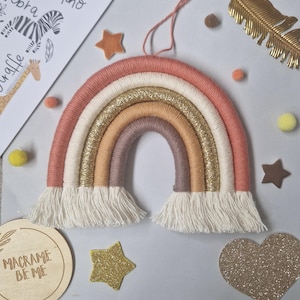 May include: A macrame rainbow in peach, cream, gold, and brown, with a fringe. Decorative stars, a heart, and a feather are placed around the rainbow. A drawing of a zebra and giraffe is in the background.