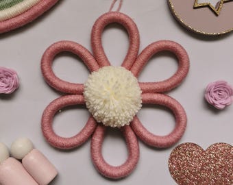 May include: A pink yarn flower with a cream-colored pom-pom center. The flower is surrounded by other decorative items, including a rainbow, a star, a heart, and small pink roses. The items are arranged on a light gray surface.
