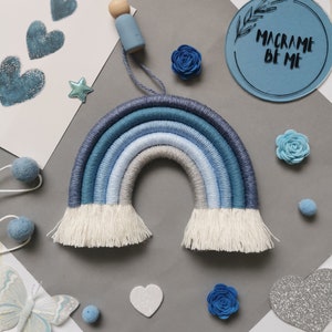 May include: A blue and white macrame rainbow wall hanging with white fringe. The rainbow is made of yarn and is hanging on a grey background. The text "Macrame Be Me" is visible in the top right corner of the image.