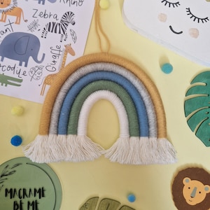 May include: A colourful macrame rainbow in shades of yellow, grey, blue, green, and white, with a fringe at the bottom. The rainbow is hanging on a yellow surface, with a decorative lion, a leaf, and a sign that says "Macrame Be Me".