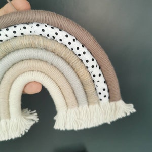 May include: A rainbow-shaped wall hanging made of yarn in various shades of beige and white. The rainbow has a white ribbon with black polka dots woven into the top layer. The bottom of the rainbow has white yarn fringe.