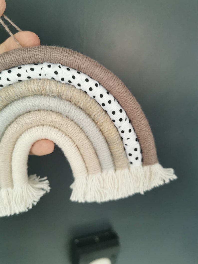 May include: A rainbow-shaped wall hanging made of yarn in various shades of beige and white. The rainbow has a white ribbon with black polka dots woven into the top layer. The bottom of the rainbow has white yarn fringe.