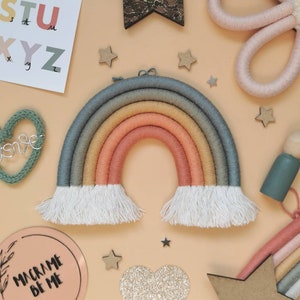 May include: A macrame rainbow wall hanging in pastel shades. The rainbow features blue, yellow, and peach colours, with white fringe. Wooden stars and a heart-shaped decoration surround the rainbow, with the text "Macrame Be Me".