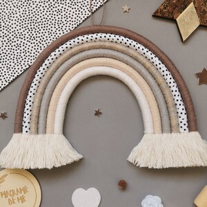 May include: A beige and brown macrame rainbow wall hanging with white fringe. The rainbow is made of multiple strands of yarn and has a polka dot pattern on the outer layer.