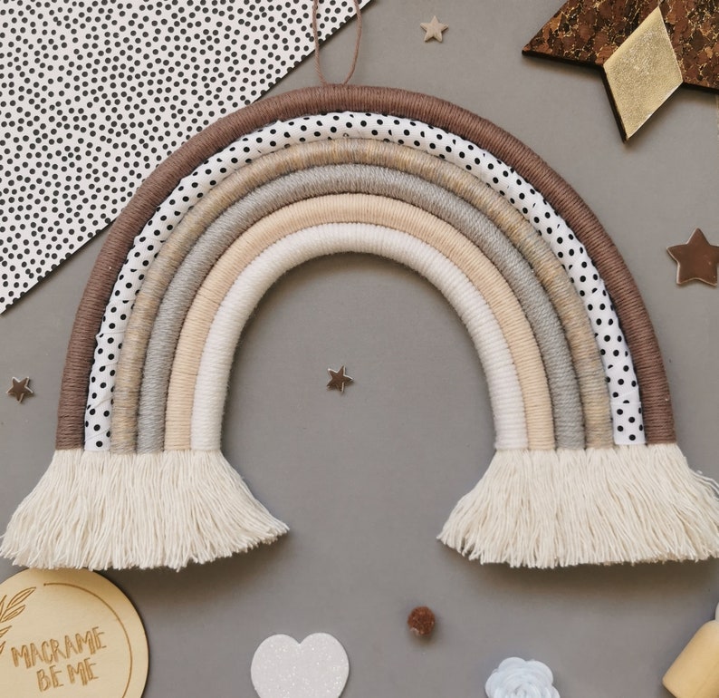 May include: A beige and brown macrame rainbow wall hanging with white fringe. The rainbow is made of multiple strands of yarn and has a polka dot pattern on the outer layer.