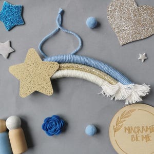 May include: A handcrafted macrame rainbow in shades of blue, gold, and white, accompanied by a gold star and a blue glitter star. Other elements include a blue felt flower, a wooden heart, and a wooden sign that says "Macrame Be Me".