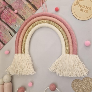 May include: A handmade macrame rainbow wall hanging in shades of pink, gold, and cream. The rainbow features a glittery gold layer and fringed ends. The background includes pink pom-poms and a wooden disc with the text "MACRAME BE ME".