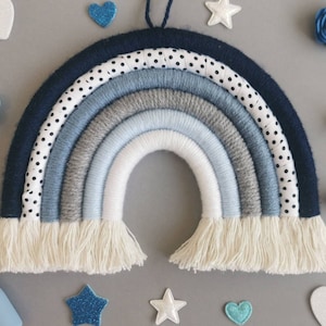 May include: A handcrafted rainbow wall decoration in blues, grey, and white. The rainbow features a navy blue outer layer, a polka dot fabric layer, and various shades of blue and grey yarn. The bottom has a fringe. Decorative stars, hearts, and flowers surround the rainbow.