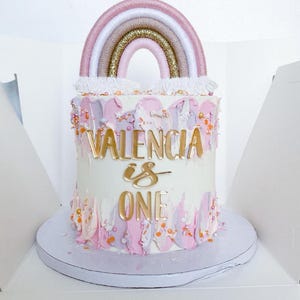 May include: A white cake with pink and purple frosting decorated with gold sprinkles and a rainbow yarn topper. The cake is decorated with gold letters that say "VALENCIA is ONE".