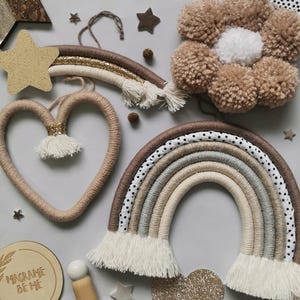 May include: A set of macrame wall hangings in neutral colors, including a rainbow, a heart, and a flower. The rainbow has a polka dot pattern and white fringe. The heart is made of natural twine. The flower is made of brown yarn.