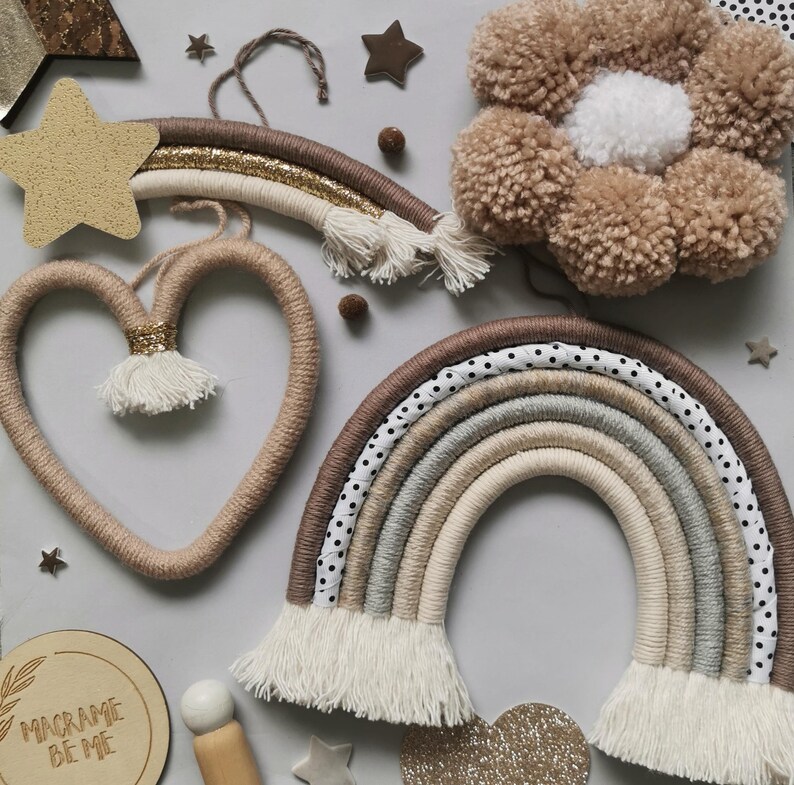 May include: A set of macrame wall hangings in neutral colors, including a rainbow, a heart, and a flower. The rainbow has a polka dot pattern and white fringe. The heart is made of natural twine. The flower is made of brown yarn.
