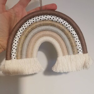 May include: A rainbow wall hanging made of woven rope and fabric. The rainbow is in shades of brown, beige, and white, with a black and white polka dot pattern on the outer layer. The rainbow has a fringe at the bottom.