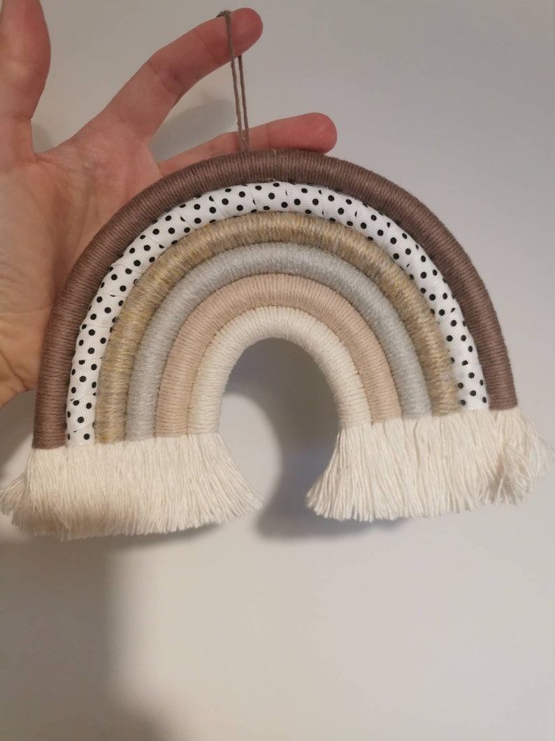 May include: A rainbow wall hanging made of woven rope and fabric. The rainbow is in shades of brown, beige, and white, with a black and white polka dot pattern on the outer layer. The rainbow has a fringe at the bottom.