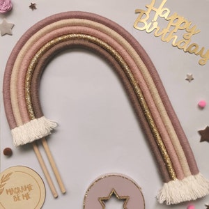 May include: A pink, beige, and gold macrame rainbow cake topper with white fringe. The topper is on two wooden sticks and has the text "Happy Birthday" in gold letters.
