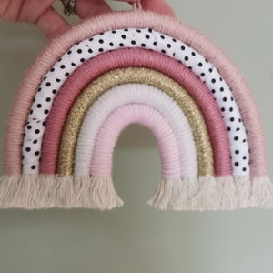 Pode incluir: Enfeite de parede de arco-&iacute;ris feito &agrave; m&atilde;o em tons de rosa, branco e dourado. O arco-&iacute;ris apresenta uma camada de tecido de bolinhas e uma franja na parte inferior. Pendurado por uma corda rosa.