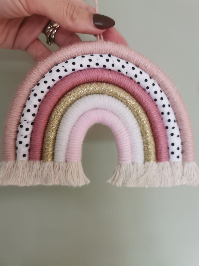 Pode incluir: Enfeite de parede de arco-&iacute;ris feito &agrave; m&atilde;o em tons de rosa, branco e dourado. O arco-&iacute;ris apresenta uma camada de tecido de bolinhas e uma franja na parte inferior. Pendurado por uma corda rosa.