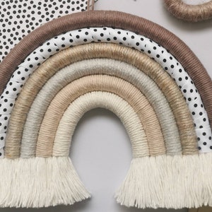May include: A beige and brown macrame rainbow wall hanging with white fringe. The rainbow is made of multiple strands of yarn and has a white and black polka dot ribbon on the outer edge.