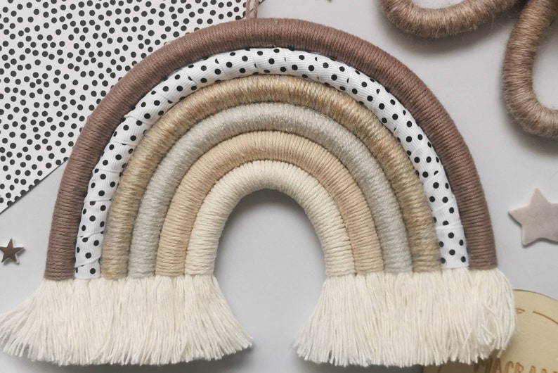May include: A beige and brown macrame rainbow wall hanging with white fringe. The rainbow is made of multiple strands of yarn and has a white and black polka dot ribbon on the outer edge.