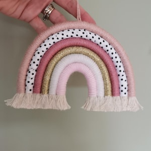 Pode incluir: Um arco-&iacute;ris decorativo feito &agrave; m&atilde;o em tons de rosa, dourado e branco, com um detalhe em tecido de bolinhas. O arco-&iacute;ris tem uma textura tecida e uma franja na parte inferior. Uma argola de corda &eacute; presa para pendurar.