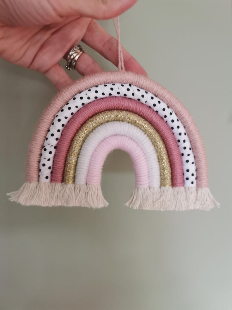 Pode incluir: Um arco-&iacute;ris decorativo feito &agrave; m&atilde;o em tons de rosa, dourado e branco, com um detalhe em tecido de bolinhas. O arco-&iacute;ris tem uma textura tecida e uma franja na parte inferior. Uma argola de corda &eacute; presa para pendurar.