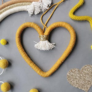 May include: Mustard yellow heart-shaped wall decoration with a gold glitter accent and white tassel. A small rainbow and yellow pom-poms are also visible. This handmade item is perfect for adding a touch of whimsy to any room.