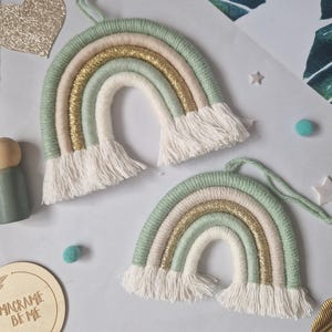 May include: Two handmade macrame rainbow wall hangings featuring wrapped yarn in sage green, cream, and shimmering gold thread. Each arch ends with white cotton fringe and includes a matching yarn loop at the top for hanging purposes.