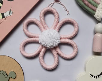 May include: A pink, handmade flower wall hanging with a white pom-pom center. The flower is made of looped yarn and has a small red pom-pom at the top for hanging. The craft item is on a white surface.
