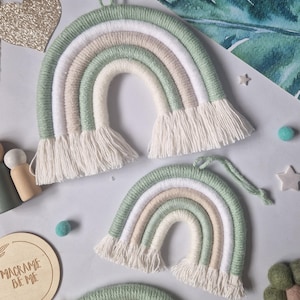 May include: Two decorative wall hangings shaped like rainbows, crafted from wrapped yarn in shades of sage green, white, and beige. Each rainbow features a soft, fringed edge at the bottom and a small loop at the top for hanging purposes.