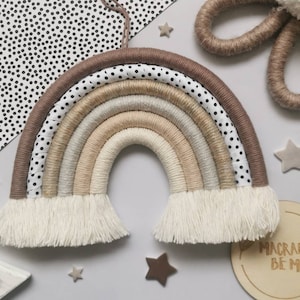 May include: A macrame rainbow wall hanging with a brown, beige, and white color scheme. The rainbow has a white fringe and is made with a variety of textures. The rainbow is hanging on a white wall with black polka dots. The text "Macrame Be Me" is visible on a wooden sign in the bottom right corner of the image.