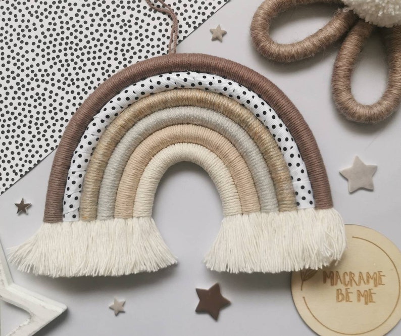 May include: A macrame rainbow wall hanging with a brown, beige, and white color scheme. The rainbow has a white fringe and is made with a variety of textures. The rainbow is hanging on a white wall with black polka dots. The text "Macrame Be Me" is visible on a wooden sign in the bottom right corner of the image.